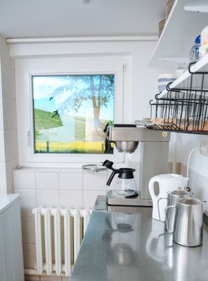 Küchenzeile mit Kaffeemaschine, Wasserkocher und Kannen auf einer Edelstahl-Arbeitsfläche vor einem Fenster