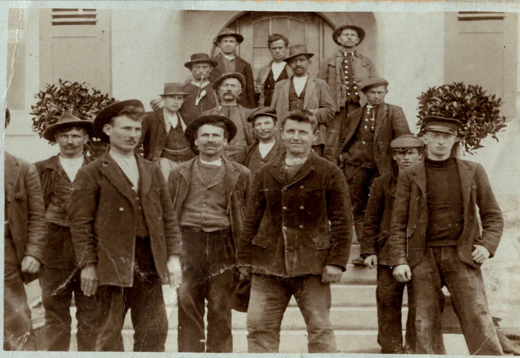 Gruppenfoto von Zimmerleuten auf einer Eingangstreppe, zwei von ihnen mit großen Laubgebinden
