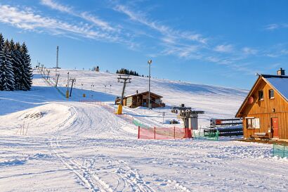 Verschneiter Skihang mit Schlepplift und Holzhütten unter blauem Himmel