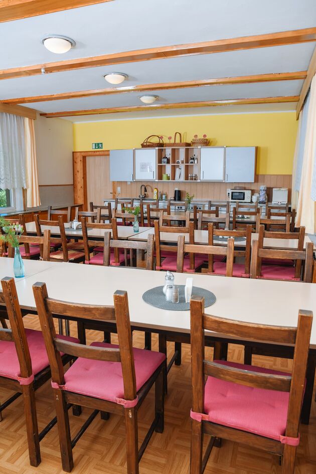 Gemeinschaftsraum mit langen Tischen, Holzstühlen mit rosa Sitzkissen und Küchenzeile an der Stirnwand