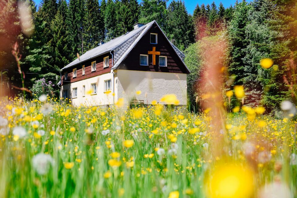 Haus mit dunkler Holzverkleidung und Holzkreuz am Giebel hinter einer blühenden Wiese, umgeben von Fichten