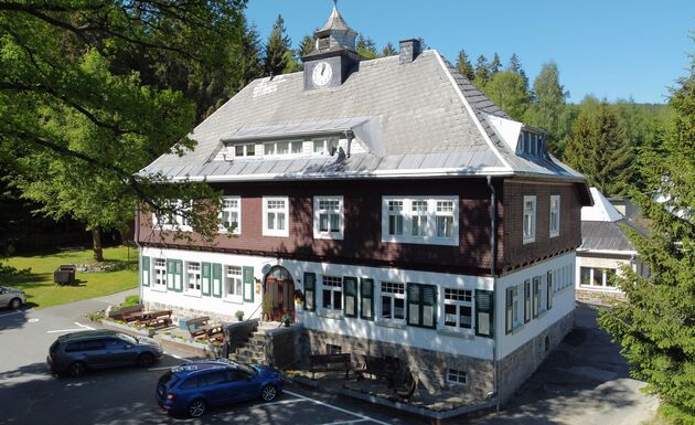 Luftbild des Haupthauses mit Uhrtürmchen und grünen Fensterläden, davor Parkplatz und Sitzbänke